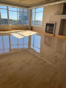 A large, open room with light-brown hardwood floors. The windows look out over a city.